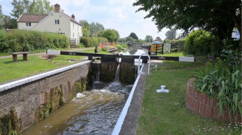 A canal is seen stopped by a lock. There is grass next to the canal and some houses.