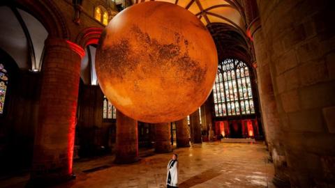 A giant representation of Mars suspended in the roof of Gloucester Cathedral with a person standing below looking up