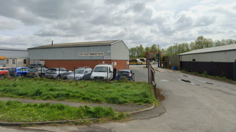A small industrial estate with a low, grey warehouse labelled “Lugg Valley Primrose Travel.” Several parked cars line the fenced front area. To the right, a narrow road leads past another warehouse, with a couple of buses visible in the background.