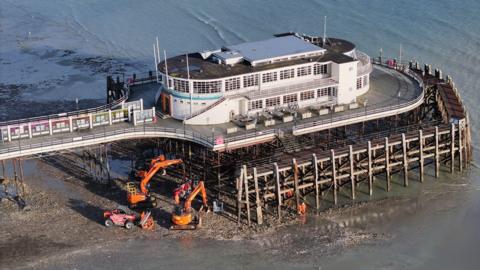 An aerial shot of orange diggers working at the base of the pier, by the support poles, during low tide. 