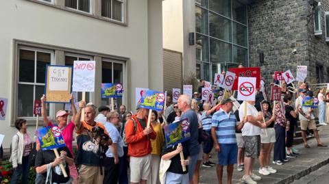 The image shows protesters standing outside Guernsey's Royal Court. There is an assortment of men and women holding placards with anti-GST signs on them. They are all standing on the pavement next to a road. 