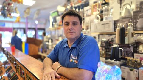 Michael Jennings is standing behind a counter. Behind him we can see there are a wide range of goods, in particular kettles.