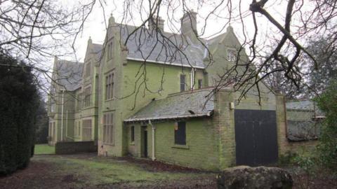 A side view of a large, boarded-up Victorian building. The weather is grey and overcast.
