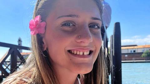Juliette, smiling at the camera on a sunny day, is wearing her long hair down, sunglasses on her head and a pink flower tucked above her ear. She is sitting by the water
