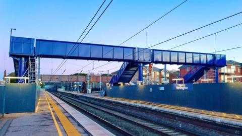 A blue footbridge connecting station platforms. There is a sign on blue hoarding saying "Welcome to Harlington". It appears to be twilight outside.