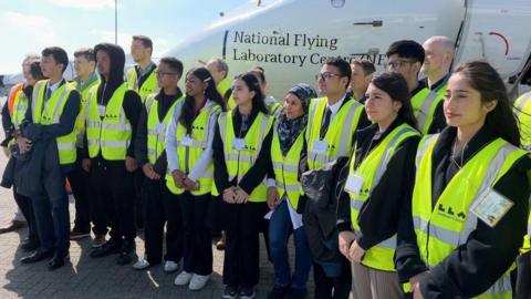 Students from Luton wearing high-vis jackets pose for a photo in front of an aircraft - the National Flying Laboratory Centre. 