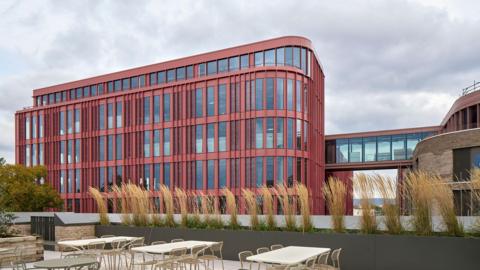The Forum in Gloucester viewed from a rooftop with chairs and tables. The Forum is a large, modern building with red coloured cladding and a glass walkway which connects it to another part of the building, which is brown brick.