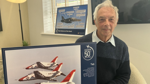 A man with white hair is sat on a cream chair, wearing a blue jumper with a white collar shirt underneath. He is holding a picture of red and white Hawk jets.