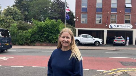 A blonde wearing a blue crew-necked jumper stood alongside a road with a lamppost in the background with an England and an Australian flag flying