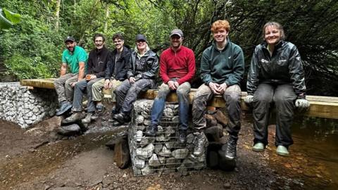 Volunteers sitting side by side along a new footbridge made of railway sleepers and stone stanchions