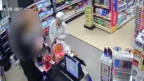 CCTV of a woman being served at a counter. The person behind the till has their face blurred. They are both standing in what looks like a small shop.