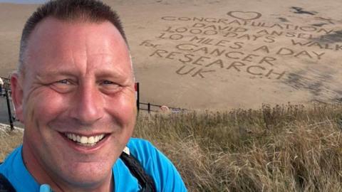 David Ansell smiling into the camera. He has short, brown hair which is grey on the sides. He is wearing a bright blue top. There is a beach behind him and words have been written into the sand along with a heart. It reads: 'Congratulations David Ansell 1000 Days walking 10 miles a day for Cancer Research UK.'