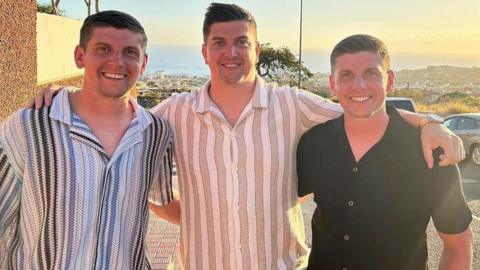 Joel, Mitchell and Curtis on holiday wearing short sleeved shirts and smiling at the camera. The three all have short dark hair and there is a coast in the background.