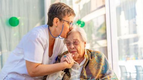 A nurse in a white uniform cares for an elderly woman, who is leaning towards her and has a scarf around her shoulders.