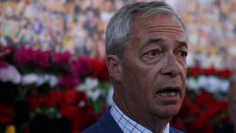 Nigel Farage speaks in north London. He is pictured with his mouth open as he speaks, wearing a blue suit and checked shirt.