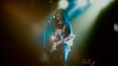 A man with long black hair singing into a microphone and playing a guitar. He's wearing leather trousers and a black top and lights are shining down on him