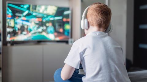A boy with ginger hair is sitting down with his back to the camera, playing a video game on a large tv in the background. He has white headphones on and is wearing a white t-shirt and blue jeans.