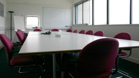 An empty meeting room with a white table surrounded by purple office chairs. There is a window to the right of the room and a whiteboard and flip chart at the far end.