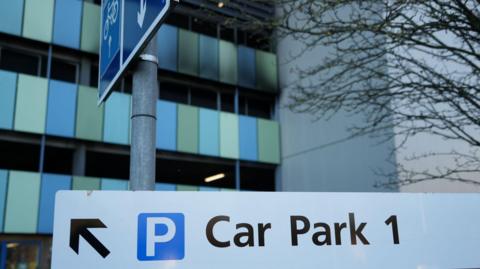 Car Park 1 at Addenbrooke's Hospital in Cambridge. It is a multi-storey car park with blue and green panels on the exterior. On part of the outside there is smoke damage and the once blue and green panels are black following a fire. In the foreground of the picture there is a white sign with an arrow pointing to the left and writing which says, Car Park 1.