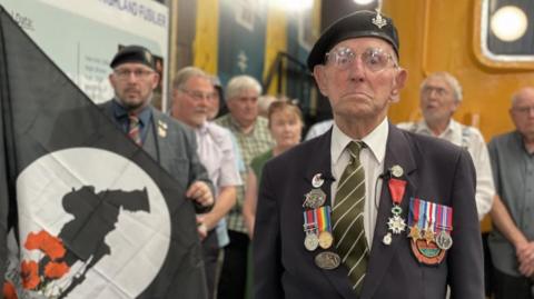 A old man wearing military medals wears a sombre expression miles as he stands in front of a group of people, a commemorative flag and a railway locomotive.