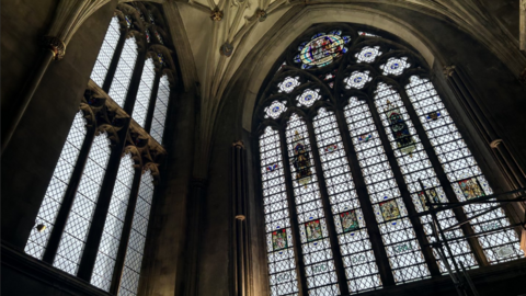 The inside of Bristol Cathedral showing the stained-glass Colston's window next to a plain window to its left. 
