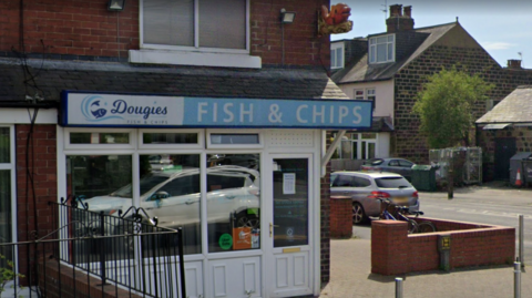 A fish and chip shop on the corner of a high street. It has white frontage and a blue sign above the door. There are cars parked nearby and a bike leaning against the wall.