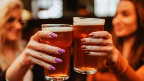 File photo showing two full pint glasses being held by one hand with purple nail varnish and another with red nail varnish. Two smiling women, out of focus, are visible behind.