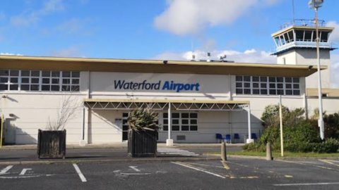 the image shows the exterior of Waterford airport in the Republic of Ireland. It is a white building with a high tower on the right of the picture. The words 'waterford airport' are written in blue above the entrance
