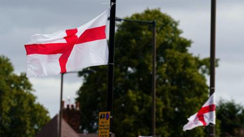 Two red and white St George's flags fly in the wind, having been tied to lampposts on a residential street.