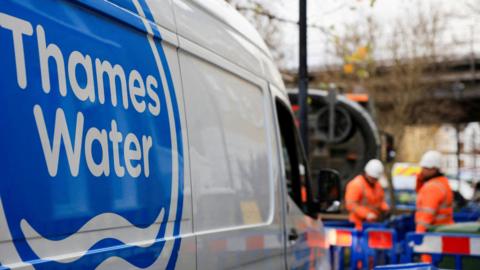 A shot of a Thames Water van. In the background there are two men wearing hi-vis clothing and hard hats and standing in an area that is boarded off by blue fences. 