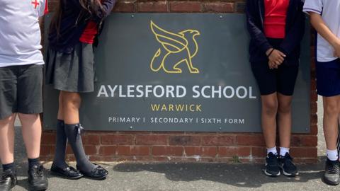 a brick wall with the sign of a school that says Aylesford School Warwick. Children can be seen standing around it in School uniform