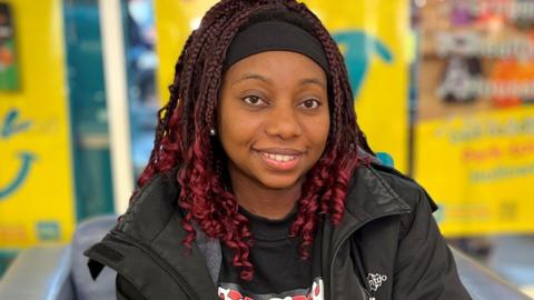 Nana is smiling into the camera and has dark red braided hair down to her shoulders and is wearing a black jacket with a small white logo at the top left on front of her jacket. Behind her are blurred yellow posters in a shop window.