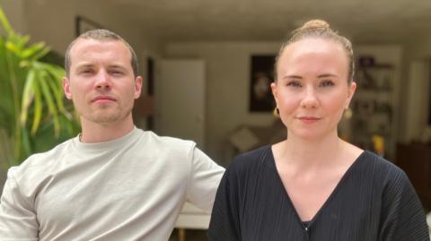 A man and woman sitting together. The man wears a pale T Shirt and the woman a black V neck T Shirt and they are sitting in a living room with plants and pictures on the wall.