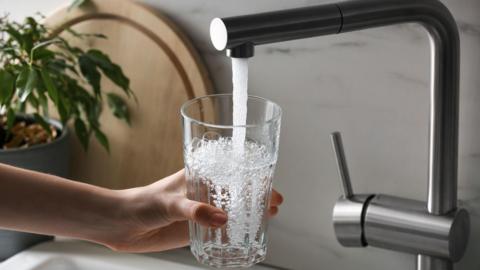 A hand holding a glass over a sink, water pouring into a glass from a silver abstract tap with a chopping board in background