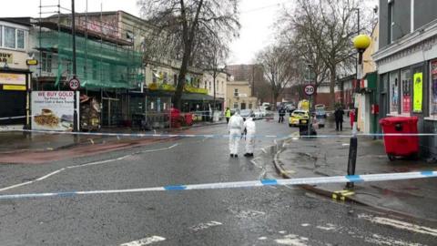 Two detectives in forensic suits look at the floor on a street behind police tape. There are a number of shops and a pub on the street, and it is a 
