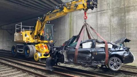 A yellow cherry-picker winches a smashed black BMW off railway tracks.