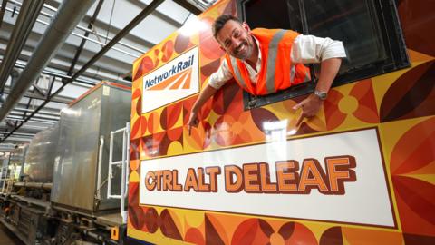 A man leans out the window of a train and points at the name painted on the side of it. He is wearing an orange high-vis jacket and the train is painted with orange and green leaves.