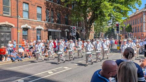 Members of a parade are marching down the streets of Belfast's Bedford Street. All are wearing white and carrying drums as people watch from the pavements. 