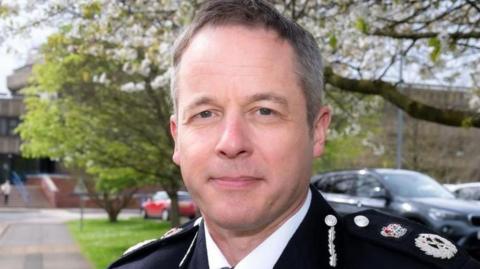 Paul has short receding grey hair and hazel-blue eyes. He is wearing a police uniform consisting of black suit jacket with badges on the lapel, over a white shirt