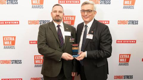 David is standing on the left and is wearing a dark green suit jacket, white shirt and blue and red tie. He has brown hair and is holding onto one side of a glass trophy. On the right is Admiral Sir Philip Jones who has silver white hair, and is wearing a white shirt, black suit jacket and black tie. He is holding on to the other side of the glass trophy.