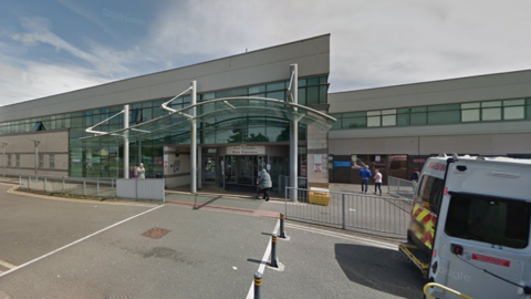 The front entrance to Ysbyty Gwynedd Hospital. Ambulance vans can be seen on the right hand side. A side reads: Main entrance" above automatic glass doors. A person walks towards the doors. 