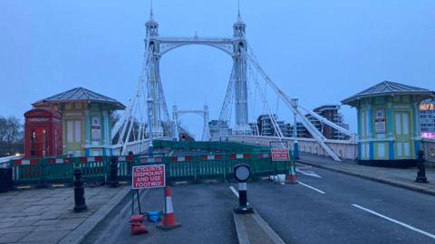 The image shows two cabled spans of the bridge, with two huts painted white with green trims on either side. There is also a red telephone box. Green barriers block the lanes for motor vehicles and a sign tells cyclists to dismount and use the footway.