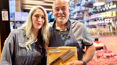 Adele Thomas has long  blonde hair and is wearing a grey dress . Geoff Hughes has a grey striped polo shirt under a traditional blue and white vertical striped butcher's apron.