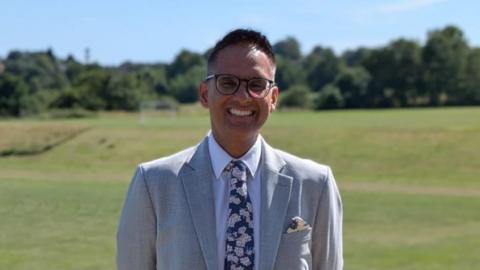 Sukhjot Dhami pictured on the school's playing field. He is wearing a grey suit, white shirt and navy floral patterned tie. He has short black hair and is also wearing think black glasses. 
