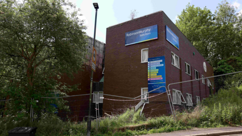 The derelict Sinfonia House in Jesmond Vale, Newcastle. The red-brick building with a flat roof is surrounded by metal fencing which is damaged. It has some open windows, with office-style white curtains.