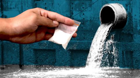 A composite graphic which shows a close-up of a hand holding a small bag of white powder - drugs - overlaid on a sewage pipe out of which a steady stream of water is emerging