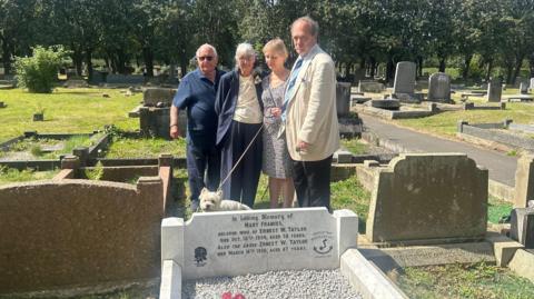 Four people stand behind a shining white grave with the words 'In Loving Memory of Mary Frances, beloved wife of Ernest W Taylor died Oct 18 1934, aged 58 years. Also the above Ernest W Taylor died March 16 1936, aged 67 years'. The rose symbol of England Rugby is on the left of the headstone while the Whitley Bay Rockcliff club badge is on the right.