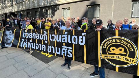 Around 60 supporters stand behind a black and yellow banner, with a message written on it in Thai in white lettering. Chansiri's face is on the side of the banner.