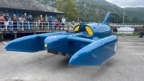 Bluebird - a jet-powered hydroplane - on display at Glenridding Pier in a car park. A number of people are looking on.