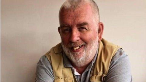 Andrew Rowland-Stuart, who died at his home on 27 May, smiling, wearing a grey t-shirt and grey waistcoat